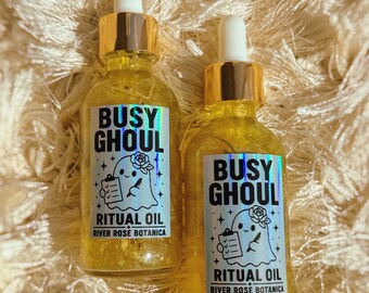 May include: Two clear glass dropper bottles filled with yellow liquid containing visible botanical pieces. Each bottle features a white label with black text reading BUSY GHOUL RITUAL OIL RIVER ROSE BOTANICA and a ghost illustration with a clipboard.