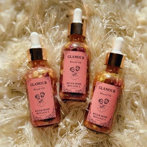 May include: Three glass bottles of "Glamour Ritual Oil" with gold droppers and pink labels. The labels read "River Rose Botanica" and feature a rose illustration. The bottles are filled with a clear liquid and floral elements.