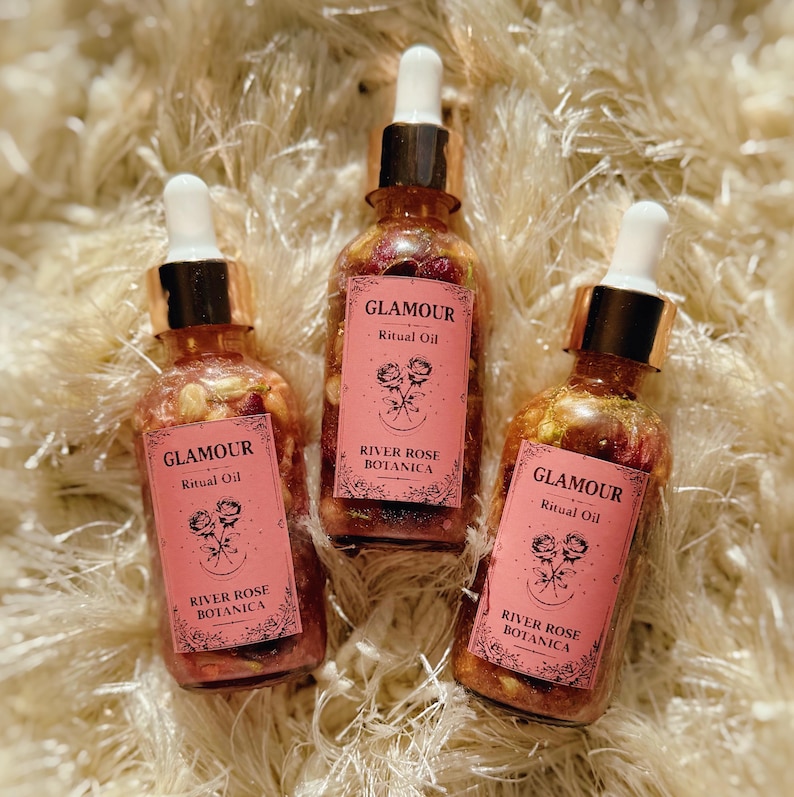 May include: Three glass bottles of "Glamour Ritual Oil" with gold droppers and pink labels. The labels read "River Rose Botanica" and feature a rose illustration. The bottles are filled with a clear liquid and floral elements.