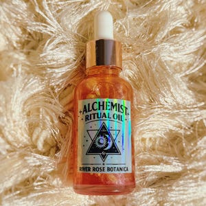 May include: A clear glass dropper bottle filled with orange liquid. The label features the text ALCHEMIST RITUAL OIL and RIVER ROSE BOTANICA with a black triangle graphic containing a sun and moon. The bottle has a metallic gold collar and white top.