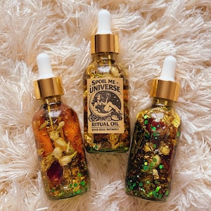 May include: Three glass ritual oil bottles with gold accents and white droppers. The centre bottle has a label that reads "Spoil Me Universe" and "Ritual Oil River Rose Botanica". The bottles contain dried flowers, glitter, and gold flakes.