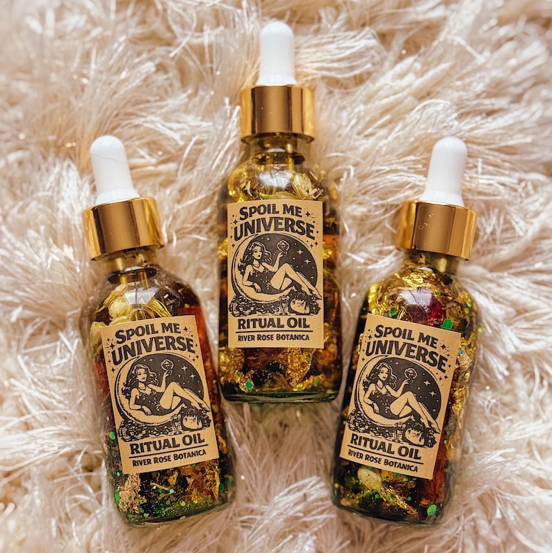 May include: Three glass bottles of ritual oil with gold droppers. Each bottle has a label with the text "Spoil Me Universe" and "Ritual Oil". The bottles are filled with a golden liquid and decorative elements.