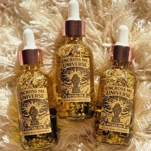May include: Three glass bottles of ritual oil with dropper tops. The bottles are filled with herbs and a golden liquid. Each bottle has a label with the text "UNCROSS ME UNIVERSE" and "RIVER ROSE BOTANICA".