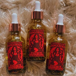 Pu&ograve; includere: Tre bottiglie di vetro di olio rituale con tappi color rame e contagocce bianchi. Ogni bottiglia ha un'etichetta rossa con la scritta "CHARMED AND DANGEROUS" e una grafica di una donna con serpenti. Il testo "RITUAL OIL RIVER ROSE BOTANICA" &egrave; anch'esso sull'etichetta.