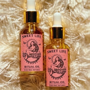 May include: Two glass bottles of ritual oil with gold-colored droppers. Each bottle has a pink label with the words "Sweet Life" and "Ritual Oil" along with an illustration of a woman. The brand name "River Rose Botanica" is also on the label.