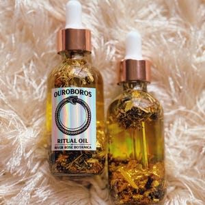 May include: Two glass bottles of ritual oil with gold flakes and botanicals. The bottles have rose gold droppers and white tops. The label reads "OUROBOROS RITUAL OIL RIVER ROSE BOTANICA" with a snake symbol.