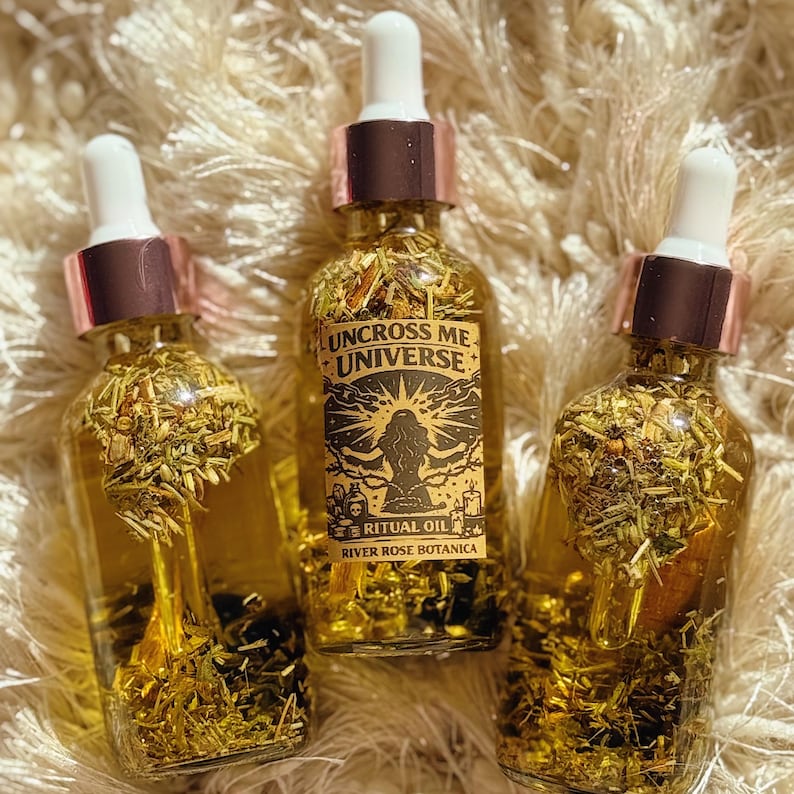 May include: Three glass bottles of ritual oil with white droppers and rose gold accents. The bottles are filled with golden liquid and herbs. The label on the center bottle reads "UNCROSS ME UNIVERSE" and "RITUAL OIL RIVER ROSE BOTANICA".