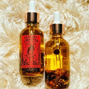 May include: Two glass bottles of ritual oil with gold-colored tops and white droppers. One bottle has a red label with the text "Destiny Swapper Block Ritual Oil" and an illustration. The other bottle contains herbs.