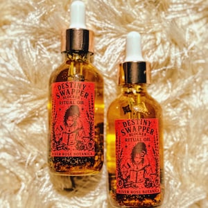 May include: Two glass bottles of ritual oil with dropper tops. Each bottle has a red label with the text "Destiny Swapper Blocker Ritual Oil" and an illustration. The brand name "River Rose Botanica" is also on the label.