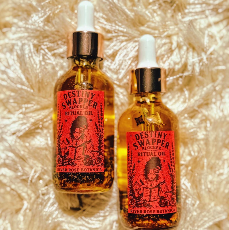 May include: Two glass bottles of ritual oil with dropper tops. Each bottle has a red label with the text "Destiny Swapper Blocker Ritual Oil" and an illustration. The brand name "River Rose Botanica" is also on the label.