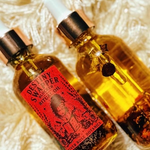 May include: Two glass bottles of ritual oil with gold-colored caps and white droppers. The bottles are filled with a golden liquid and contain botanicals. One bottle has a red label with the text "Destiny Swapper Block Ritual Oil" and an illustration.