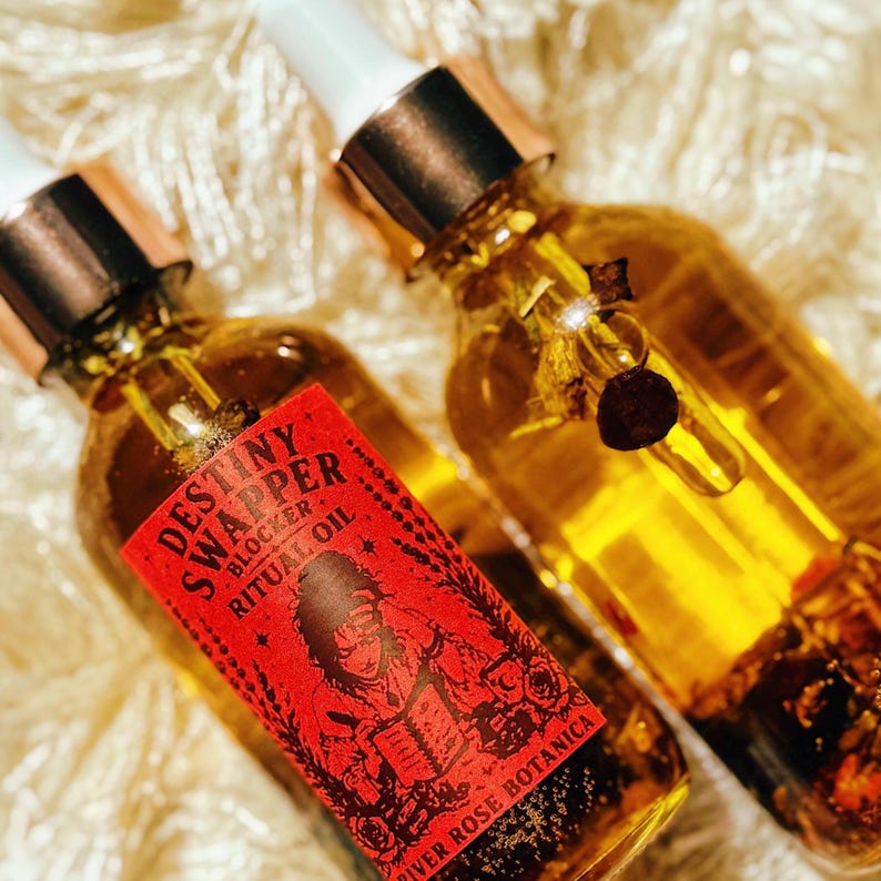 May include: Two glass bottles of ritual oil with gold-colored caps and white droppers. The bottles are filled with a golden liquid and contain botanicals. One bottle has a red label with the text "Destiny Swapper Block Ritual Oil" and an illustration.