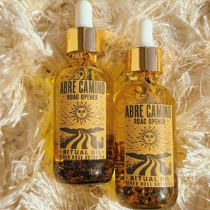 Può includere: Due flaconi contagocce in vetro trasparente riempiti con liquido dorato e pezzi botanici. Ogni flacone presenta un'etichetta color crema con testo nero che recita ABRE CAMINO ROAD OPENER RITUAL OIL RIVER ROSE BOTANICA e un sole su una strada.