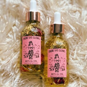 May include: Two glass bottles of ritual oil with dropper tops. The bottles have pink labels with the text "THERE SHE GLOWS" and "RITUAL OIL". The oil contains herbs and rose petals. The labels also feature an illustration of a woman.