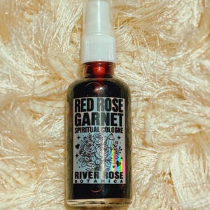 May include: A clear glass spray bottle containing a dark liquid, topped with a white plastic pump nozzle. The label features the text RED ROSE GARNET SPIRITUAL COLOGNE and RIVER ROSE BOTANICA, accompanied by a graphic illustration of roses and a crystal.