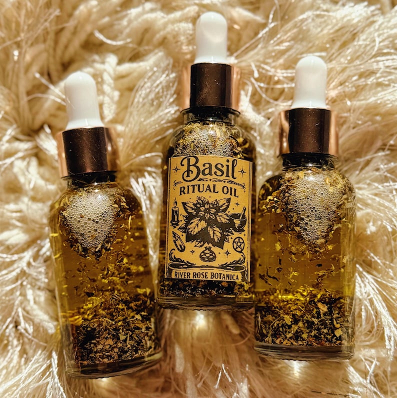 May include: Three glass bottles of Basil Ritual Oil with gold-colored liquid and herbs. Each bottle has a white dropper and a copper-colored cap. The label reads "Basil Ritual Oil" and "River Rose Botanica" with botanical illustrations.