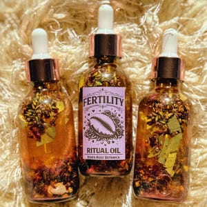 May include: Three glass bottles filled with a golden liquid and botanicals. Each bottle has a dropper, a copper-colored cap, and a label that reads "FERTILITY RITUAL OIL" with a seashell illustration. The bottles are arranged on a textured surface.