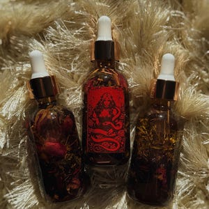 May include: Three glass bottles with dropper tops, filled with a dark liquid and botanicals. The center bottle has a red label with a woman and snake illustration. The bottles are on a textured, beige surface.