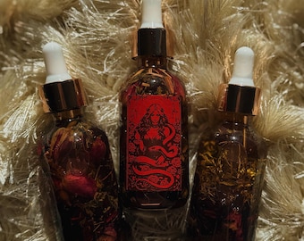 May include: Three glass bottles with dropper tops, filled with a dark liquid and botanicals. The center bottle has a red label with a woman and snake illustration. The bottles are on a textured, beige surface.