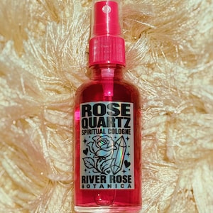 May include: A clear glass spray bottle filled with vibrant pink liquid. The label features black text reading ROSE QUARTZ SPIRITUAL COLOGNE and RIVER ROSE BOTANICA, accompanied by a graphic illustration of a rose and a crystal point.