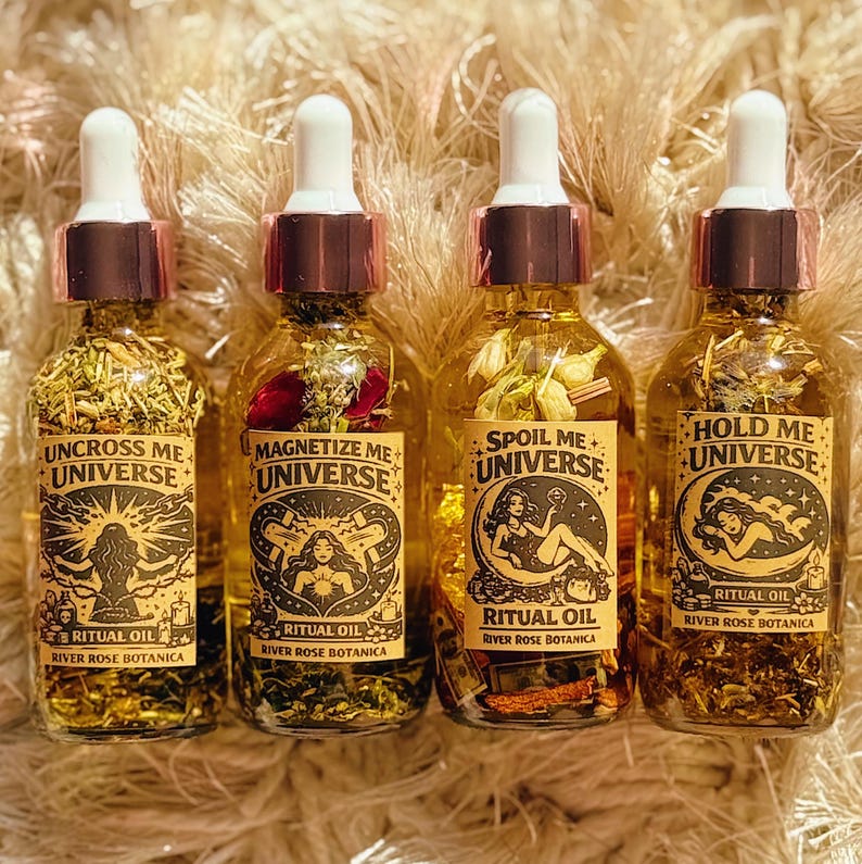 May include: Four glass bottles of ritual oil with white droppers and copper-colored caps. Each bottle has a different label with text and illustrations: "Uncross Me Universe," "Magnetize Me Universe," "Spoil Me Universe," and "Hold Me Universe."