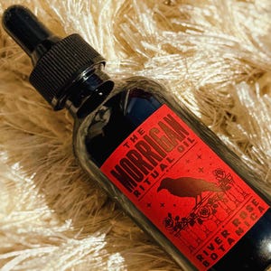May include: A dark glass dropper bottle featuring a red rectangular label with black text that reads The Morrigan Ritual Oil and River Rose Botanica. The label displays a black silhouette of a crow perched on a fence surrounded by roses and stars.