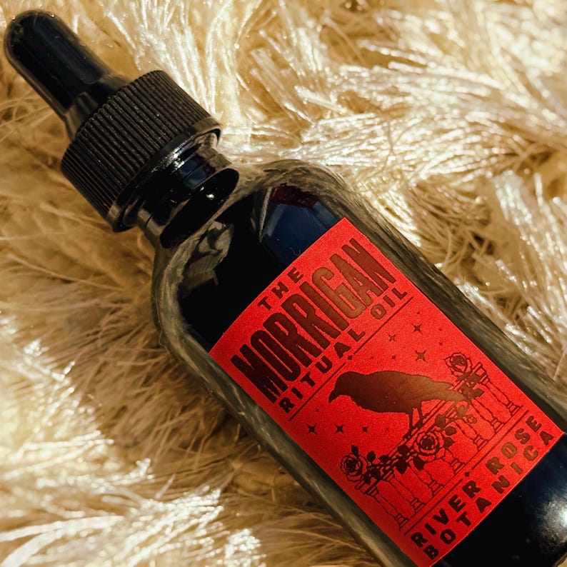 May include: A dark glass dropper bottle featuring a red rectangular label with black text that reads The Morrigan Ritual Oil and River Rose Botanica. The label displays a black silhouette of a crow perched on a fence surrounded by roses and stars.