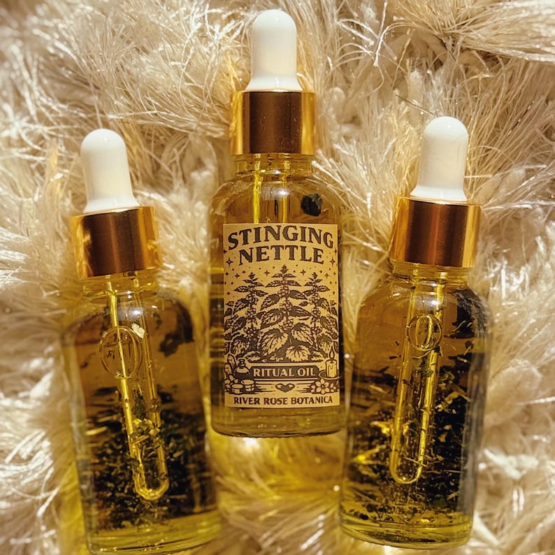 May include: Three glass dropper bottles filled with yellow liquid. The center bottle has a label that reads "Stinging Nettle Ritual Oil" with botanical illustrations. The bottles have gold-coloured caps and white droppers. The background is a fluffy, light-coloured surface.