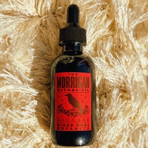 May include: A dark glass dropper bottle with a black cap, featuring a rectangular red label. The label displays the text THE MORRIGAN RITUAL OIL and RIVER ROSE BOTANICA, with a black crow illustration perched on a floral railing design.