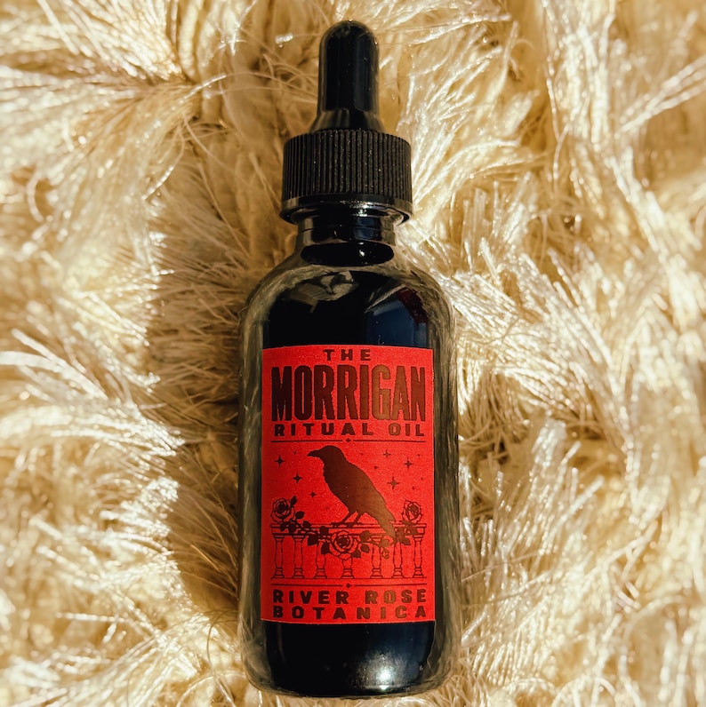 May include: A dark glass dropper bottle with a black cap, featuring a rectangular red label. The label displays the text THE MORRIGAN RITUAL OIL and RIVER ROSE BOTANICA, with a black crow illustration perched on a floral railing design.