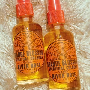 Puede incluir: Dos frascos de spray de vidrio transparente llenos de l&iacute;quido color &aacute;mbar. Cada frasco tiene una etiqueta naranja con un gr&aacute;fico de una rodaja de c&iacute;trico y el texto: Orange Blossom Spiritual Cologne, River Rose Botanica y una peque&ntilde;a flor.