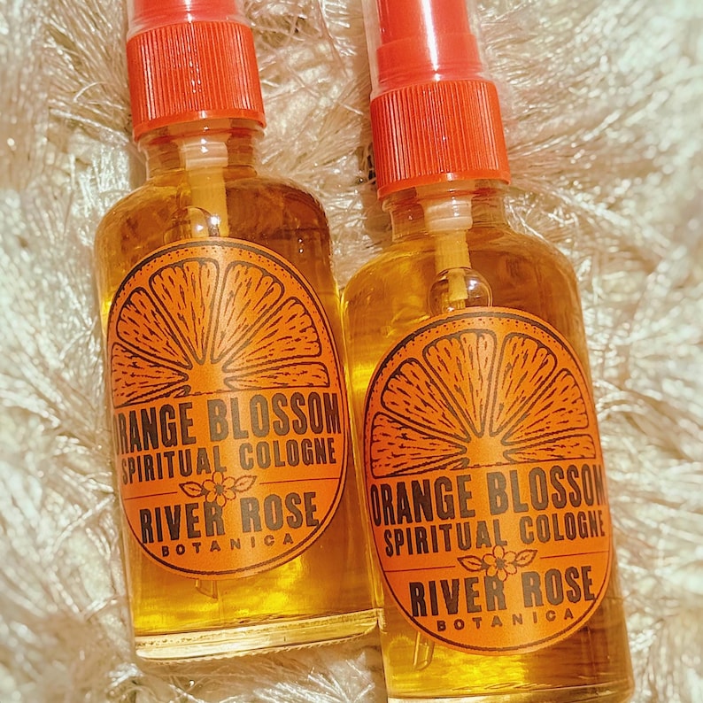 Puede incluir: Dos frascos de spray de vidrio transparente llenos de l&iacute;quido color &aacute;mbar. Cada frasco tiene una etiqueta naranja con un gr&aacute;fico de una rodaja de c&iacute;trico y el texto: Orange Blossom Spiritual Cologne, River Rose Botanica y una peque&ntilde;a flor.