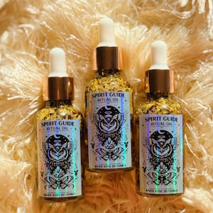 Puede incluir: Tres botellas de vidrio de Aceite Ritual Guía Espiritual con detalles dorados y cuentagotas blancos. Cada botella tiene una etiqueta con un diseño de búho y mariposa, y el texto "Spirit Guide Ritual Oil" y "River Rose Botanica". Las botellas están llenas de copos dorados.