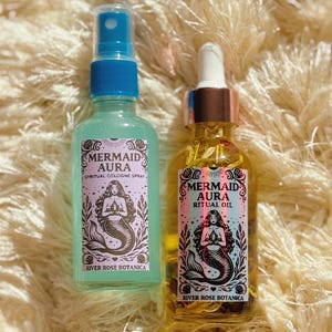 以下が含まれることがあります： 2つのMermaid Aura製品のガラス瓶。1つは明るい緑色のスプレーボトル、もう1つは金色の儀式用オイルボトルです。どちらにも人魚のイラストと「Mermaid Aura」と「River Rose Botanica」の文字が書かれたラベルが付いています。