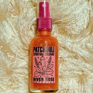 Peut inclure: Un flacon pulvérisateur en verre transparent rempli de liquide orange, avec une étiquette rose portant le texte noir PATCHOULI SPIRITUAL COLOGNE et RIVER ROSE BOTANICA, orné d'un dessin au trait noir représentant deux cristaux et des branches.