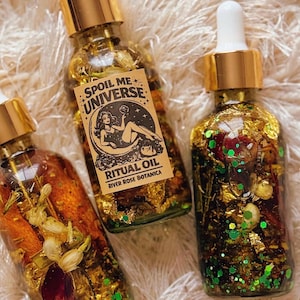 May include: Three glass bottles of ritual oil with gold lids and droppers. The bottles contain gold flakes, green glitter, and botanicals. One bottle has a label reading "SPOIL ME UNIVERSE" and "RITUAL OIL".