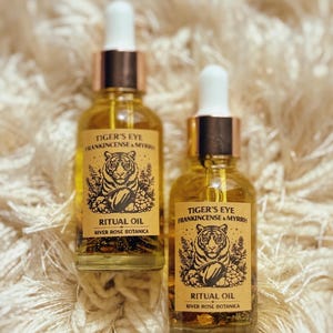 Pode incluir: Duas garrafas de vidro de Óleo Ritual Olho de Tigre com conta-gotas. Os rótulos apresentam uma ilustração de tigre e o texto "Tiger's Eye Frankincense & Myrrh" e "River Rose Botanica". O óleo é de cor âmbar.