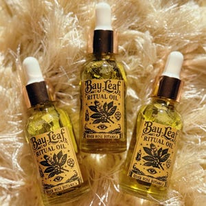 Puede incluir: Tres botellas de vidrio de Aceite Ritual de Laurel con cuentagotas blancos y detalles dorados. Cada botella tiene una etiqueta con un diseño de hoja, un ojo y el texto "Bay Leaf Ritual Oil" y "River Rose Botanica". Las botellas están llenas de un líquido amarillo.