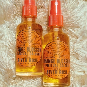 Puede incluir: Dos frascos de spray de vidrio transparente llenos de l&iacute;quido &aacute;mbar. Cada frasco tiene una etiqueta naranja con un gr&aacute;fico de una rodaja de c&iacute;trico y el texto: ORANGE BLOSSOM SPIRITUAL COLOGNE RIVER ROSE BOTANICA. Boquillas de color rojo.