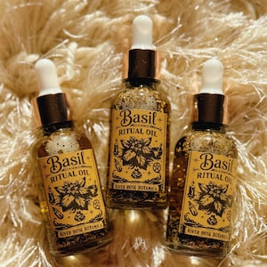 May include: Three glass bottles of Basil Ritual Oil with white droppers and gold-colored caps. Each bottle has a label with the words "Basil Ritual Oil" and "River Rose Botanica" in a decorative font. The bottles are filled with a golden liquid and herbs.
