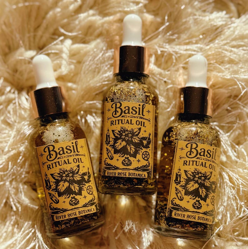 May include: Three glass bottles of Basil Ritual Oil with white droppers and gold-colored caps. Each bottle has a label with the words "Basil Ritual Oil" and "River Rose Botanica" in a decorative font. The bottles are filled with a golden liquid and herbs.