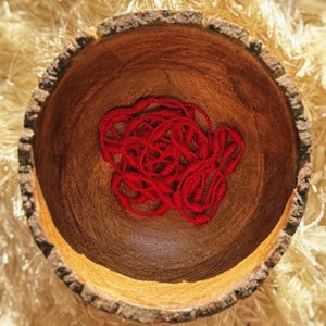 May include: A wooden bowl with a rough bark edge, filled with a tangled mass of bright red yarn. The bowl's interior is a warm brown, contrasting with the vibrant yarn. The background is a light, textured surface.