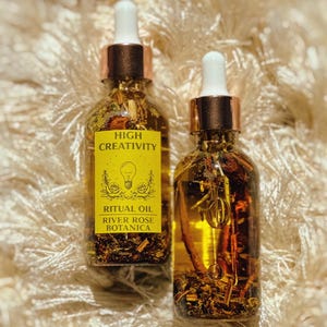 May include: Two glass bottles of ritual oil with gold-colored caps and white droppers. The bottles are filled with a golden liquid and botanical elements. One bottle has a yellow label with the text "HIGH CREATIVITY" and "RITUAL OIL RIVER ROSE BOTANICA".