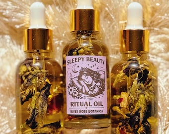May include: Three glass bottles of ritual oil with gold-colored caps and white droppers. The bottles are filled with a clear liquid and dried flowers. The center bottle has a label that reads "Sleepy Beauty Ritual Oil" with a sleeping woman illustration.