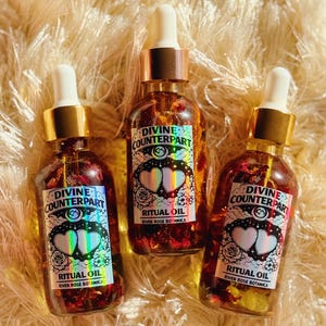 Könnte beinhalten: Drei Glasflaschen mit Ritualöl mit goldfarbenen Kappen und weißen Tropfern. Jede Flasche hat ein Etikett mit dem Text "DIVINE COUNTERPART" und "RITUAL OIL RIVER ROSE BOTANICA". Die Flaschen sind mit einer rötlichen Flüssigkeit und Blütenblättern gefüllt.