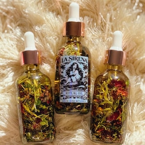 May include: Three glass bottles of ritual oil with white droppers and copper-colored collars. The bottles are filled with herbs and flowers. The center bottle has a label with a mermaid illustration and the words "LA SIRENA RITUAL OIL".