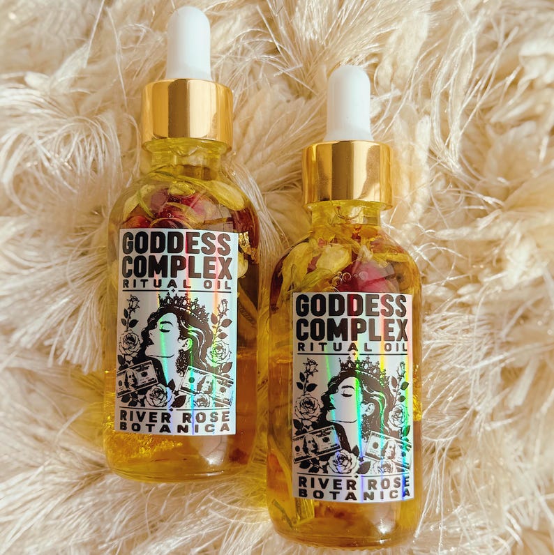 K&ouml;nnte beinhalten: Zwei klare Glastropfflaschen gef&uuml;llt mit gelbem &Ouml;l und getrockneten Bl&uuml;tenbl&auml;ttern. Jedes Etikett tr&auml;gt den Text Goddess Complex Ritual Oil River Rose Botanica, eine Grafik einer Person mit Krone sowie Abbildungen von Geldscheinen.