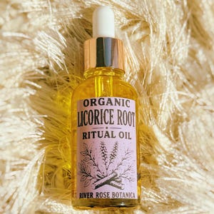 May include: A clear glass dropper bottle filled with golden yellow oil. The label is light purple with black text reading ORGANIC LICORICE ROOT RITUAL OIL and RIVER ROSE BOTANICA, featuring a botanical illustration of leaves and root segments.