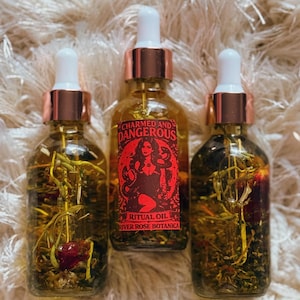 Puede incluir: Tres botellas de vidrio de aceite ritual con cuentagotas blancos y collares color cobre. La botella central tiene una etiqueta roja con una mujer y las palabras "Charmed and Dangerous". Las botellas están llenas de hierbas y flores.