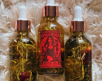 May include: Three glass bottles of ritual oil with white droppers and copper-colored collars. The center bottle has a red label with a woman and the words "Charmed and Dangerous". The bottles are filled with herbs and flowers.